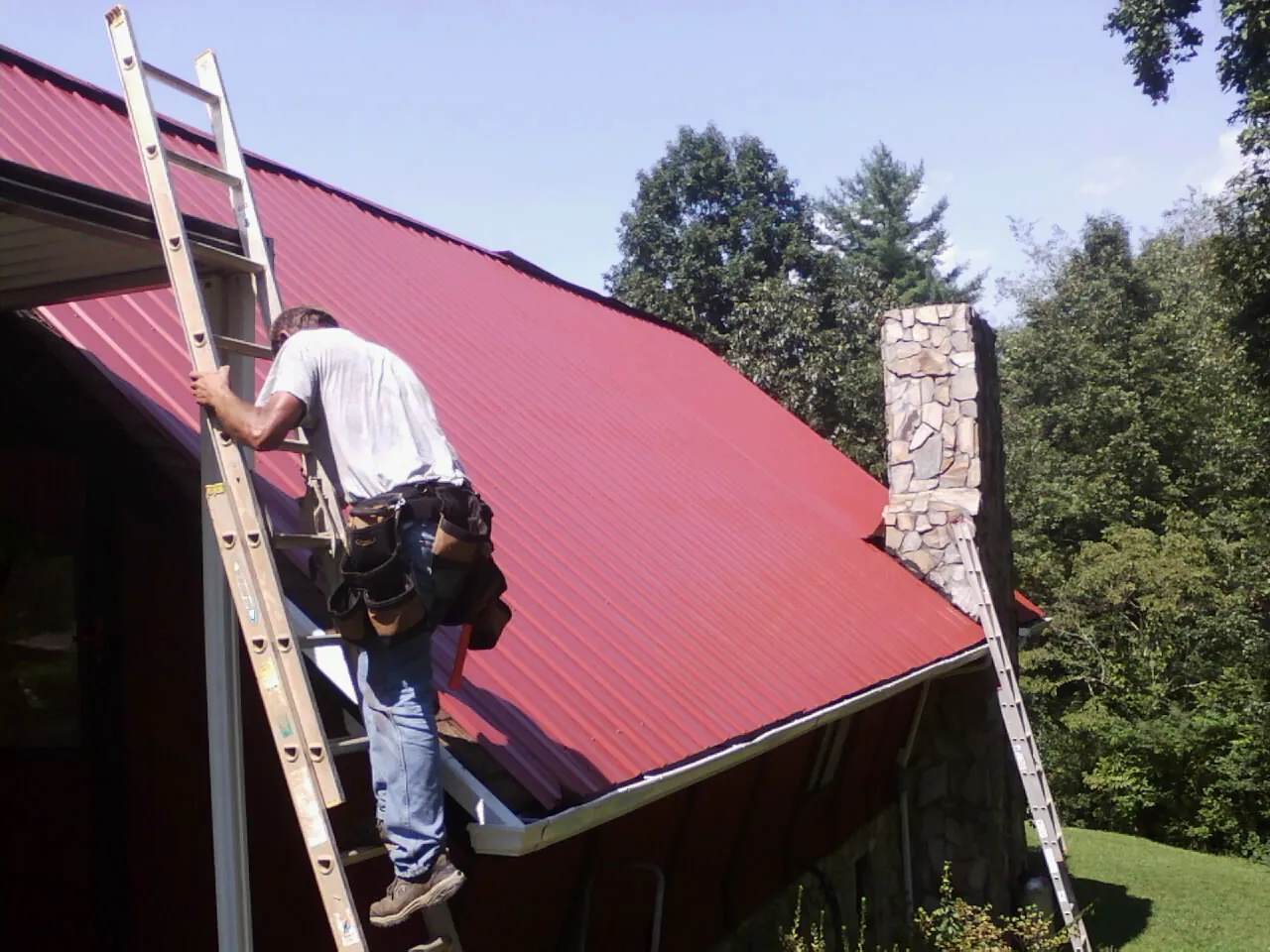 Roofer on a ladder beside a red metal roof and stone chimney — Soffit & Fascia Repair by Roofing Brothers in Broussard